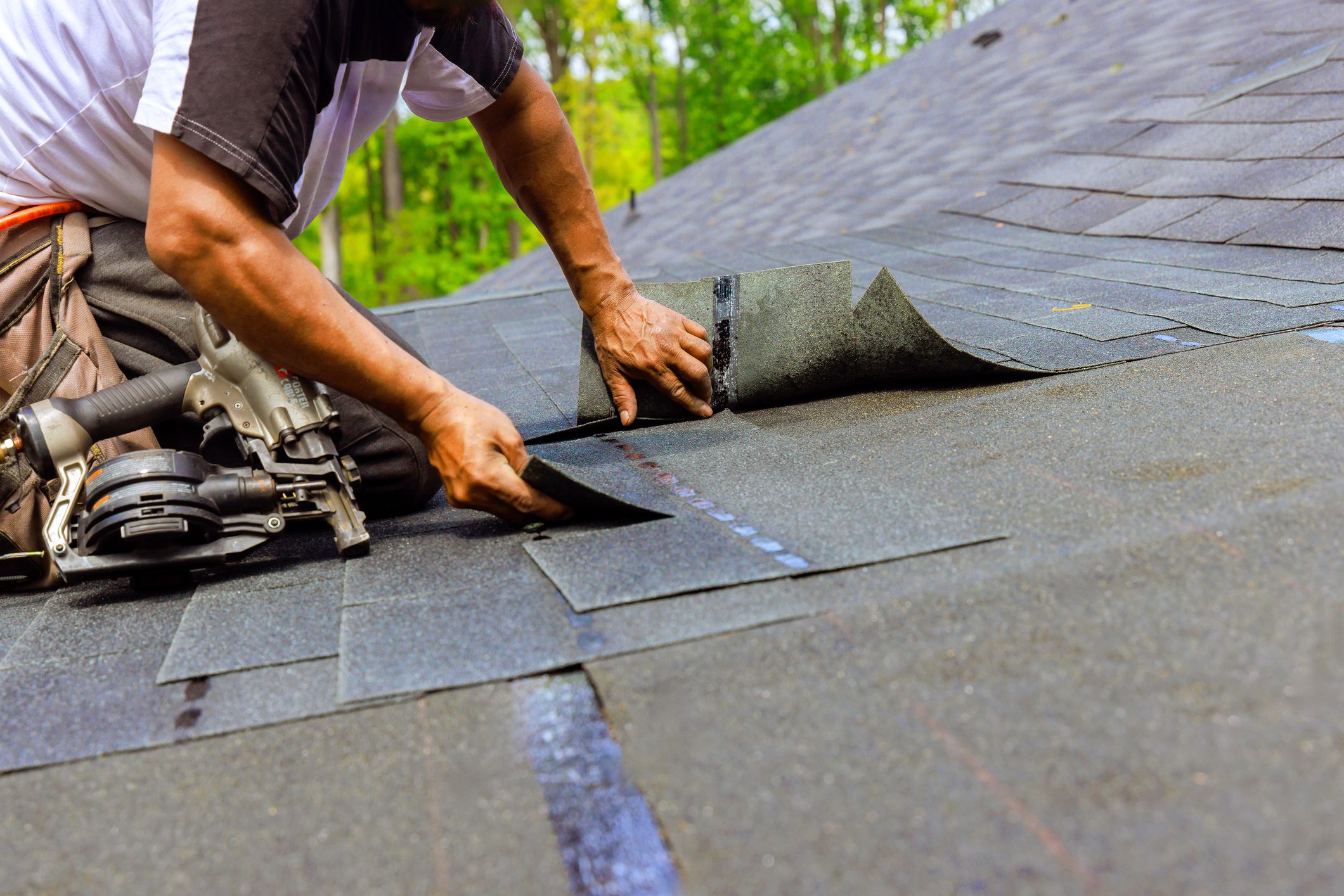 Construction worker using pneumatic nail gun to install new bitumen shingles on roof