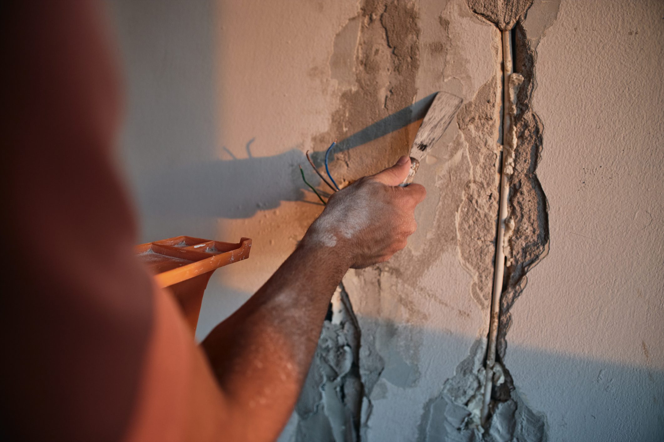 Close-up of a male worker repairing damaged plaster on a wall with a trowel. The image shows detailed texture of cracked wall surface and construction work in progress indoors with warm lighting.